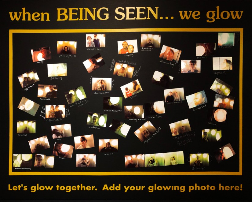 A black display board framed in gold with the gold vinyl title "when BEING SEEN... we glow" at the top and "Let's glow together. Add your glowing photo here!" at the bottom, covered with instant sticker selfies of visitors inside the Glow Chamber installation by Kingston artist Portia Po Chapman.