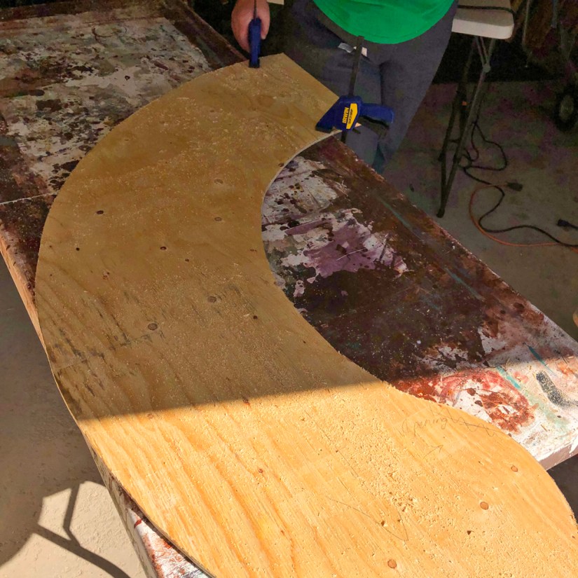 A curved, precision-cut plywood base segment clamped to a studio workbench in a wood shop.