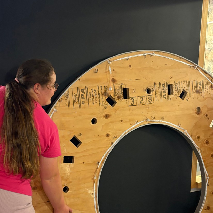 Kingston artist Portia Po Chapman supporting the circular wooden underside of the Glow Chamber's integrated plinth top as it leans against a dark gallery wall, showing precise cutouts and integrated LED tape lighting along the rim.