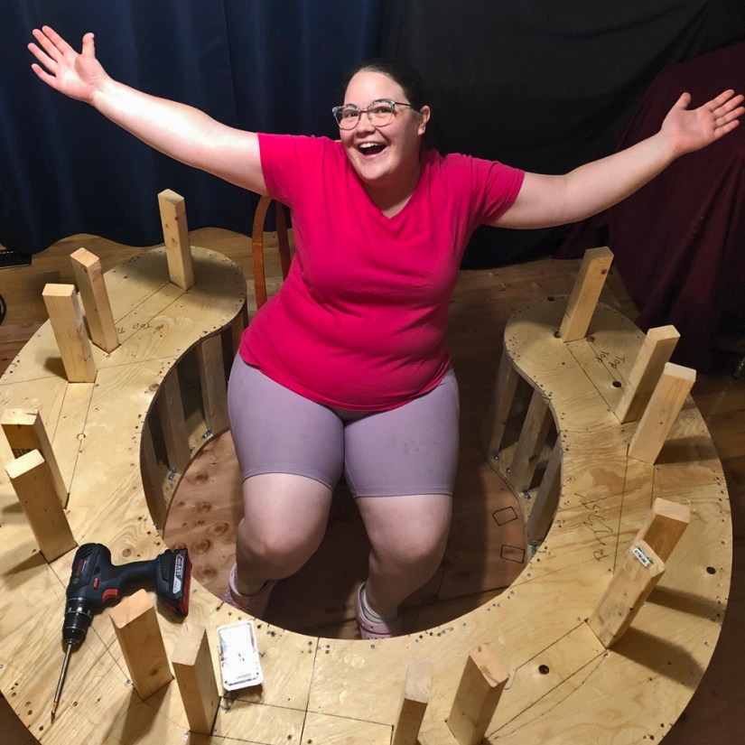 Smiling artist Portia Po Chapman sitting joyfully within the curved, partially assembled wooden integrated plinth base with vertical stud legs.