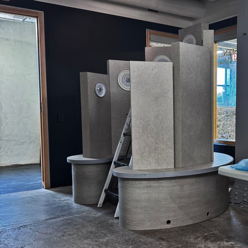 The fully assembled wooden and textured grey monolith Glow Chamber structure standing in the Pocket Gallery with backlighting fixtures mounted, showing wiring access holes along the plinth base.