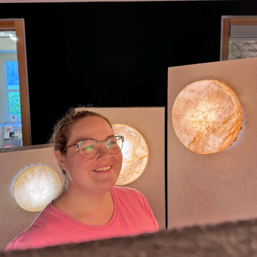 Kingston artist Portia Po Chapman smiling radiantly as she adjusts dimmer lighting behind backlit circular rawhide artworks inside the Glow Chamber installation.