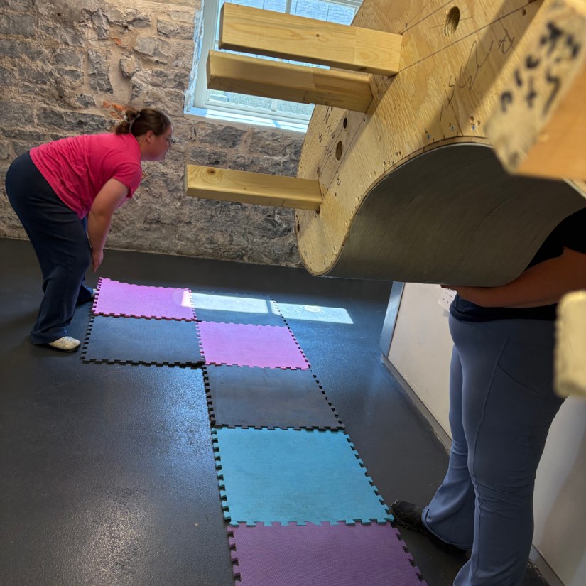 Kingston artist Po Chapman bending down in the Tett Centre hallway to position colourful interlocking foam tiles while watching for passersby before sliding the Glow Chamber plinth toward the gallery doorway.