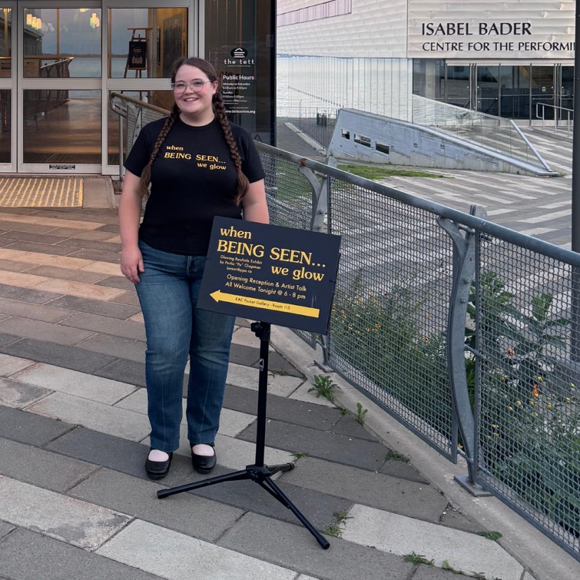 Kingston artist Portia Po Chapman standing outside the Tett Centre wearing a custom Being Seen shirt beside a directional opening reception sign pointing to the Pocket Gallery.