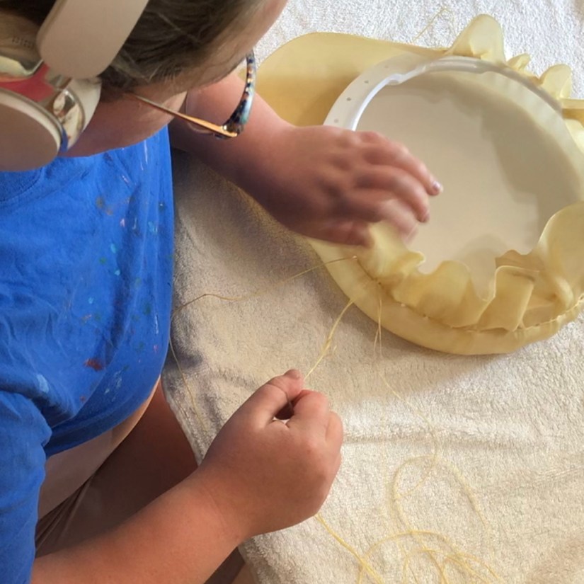 Top-down view of Kingston artist Portia Po Chapman in her studio, wearing headphones and eyeglasses, using synthetic sinew and a backstitch pattern to lace water-soaked rawhide over a pre-drilled plastic LED diffuser frame. An excessive margin of rawhide forms a ruffle, which Po intends to finger-mold and progressively trim to create a hide skirt.