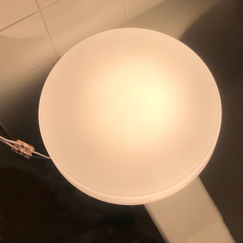Top-down view of a circular LED light fixture with an opaque plastic diffuser shade glowing softly, wired with quick-connect terminals during a studio test by Portia Po Chapman.
