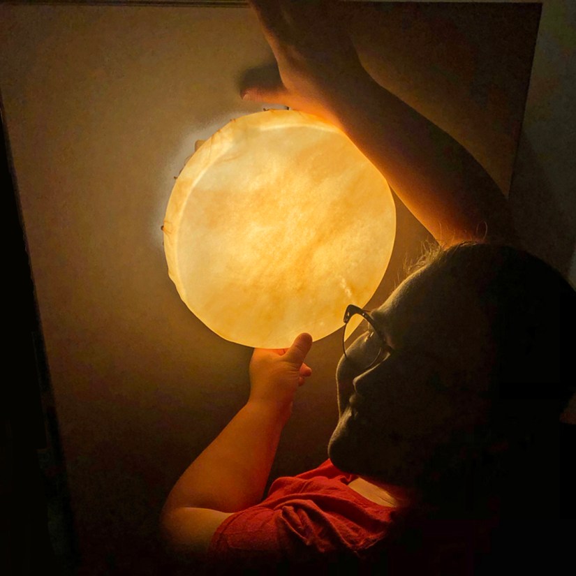 Kingston artist Portia Po Chapman smiling in a dimly lit studio as she holds an illuminated, circular rawhide vessel glowing with a warm golden sunlit light against a panel of stone-hybrid sheeting.