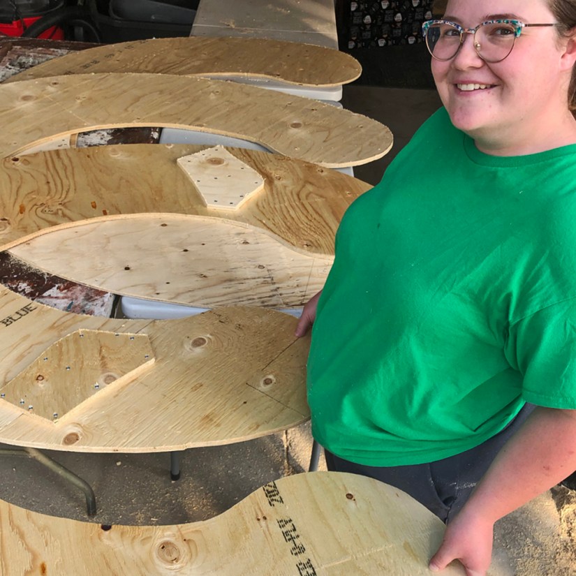 Kingston artist Portia Po Chapman in her studio wood shop standing over plywood base arcs showing temporary joining braces and offset seams.