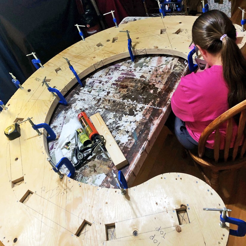 Kingston artist Portia Po Chapman seated at a workbench tracing leg placement marks onto clamped layers of a large curved plywood base.