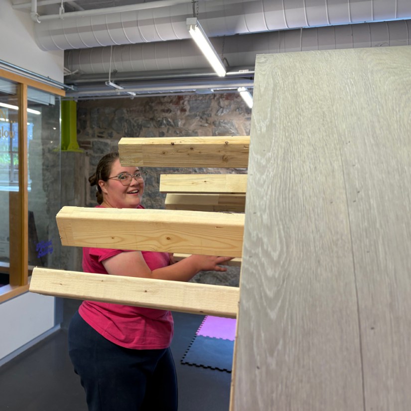 Kingston artist Po Chapman smiling as she guides the wooden Glow Chamber plinth base—wrapped in grey oak-look vinyl—through the side doorway into the Tett Centre hallway.