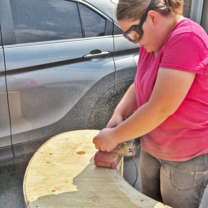 Kingston artist Portia Po Chapman using a belt sander on the flipped-up base of the wooden Glow Chamber plinth outside her studio to ensure a level fit on gallery concrete floors.