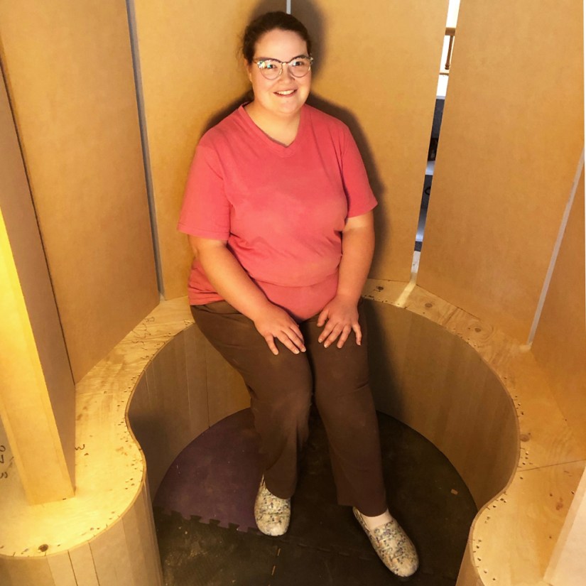Kingston artist Portia Po Chapman seated inside the semi-circular enclosure of wooden monoliths bathed in a warm, golden glow.