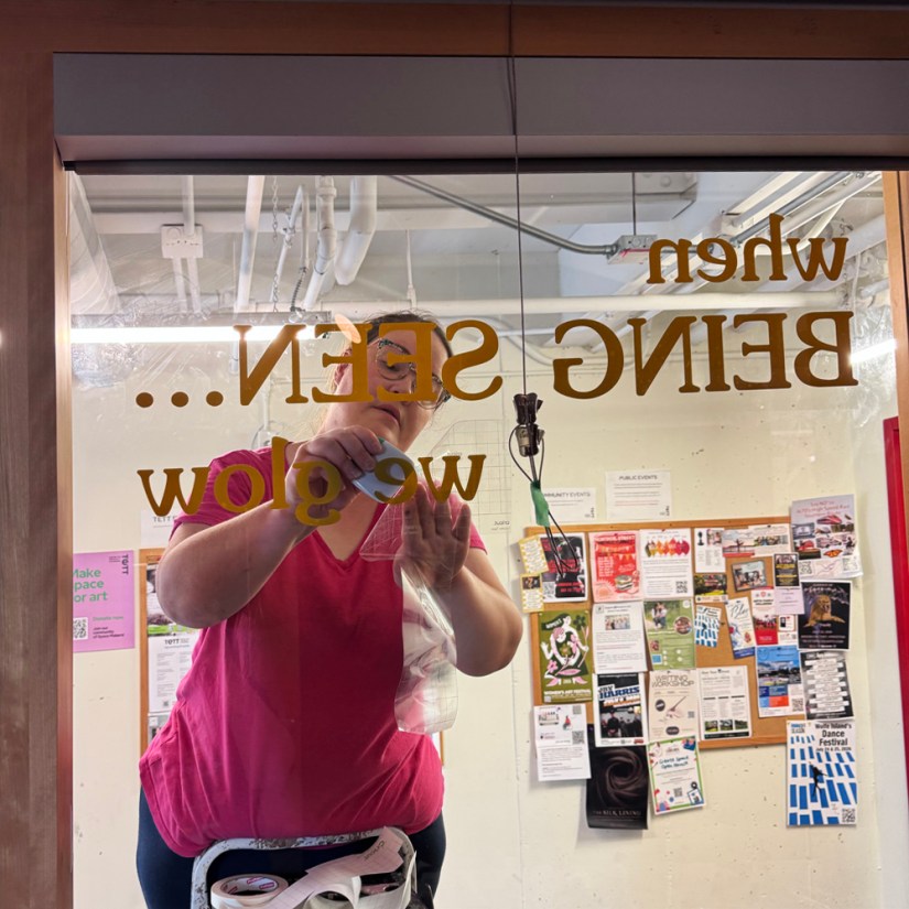 View from inside the gallery showing Kingston artist Portia Po Chapman on a stepladder applying gold vinyl typography reading "when Being Seen... we glow" onto the outer hallway window at the Tett Centre.
