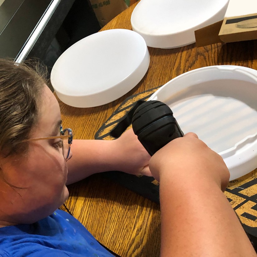 Kingston artist Portia Po Chapman drilling a hole pattern into a white plastic LED light diffuser, supported by a black sponge kneeling pad, in her studio as she prepares the backing for rawhide wrapping.