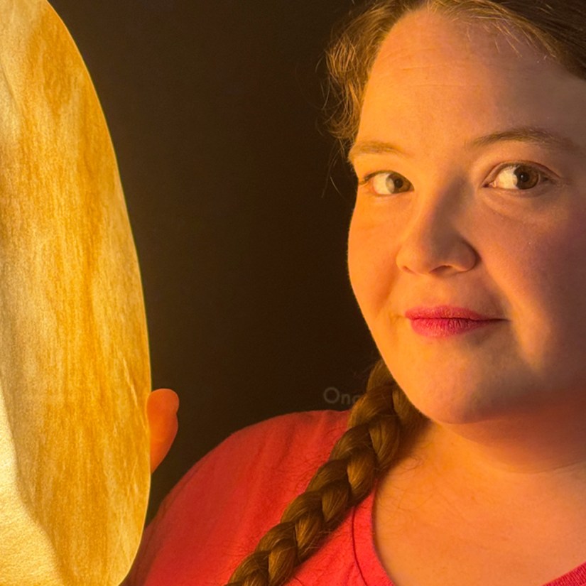 Close-up portrait of Kingston artist Portia Po Chapman in a pink top with her hair braided, illuminated by the warm golden light emitted from a handcrafted rawhide vessel she holds in the dark Pocket Gallery space