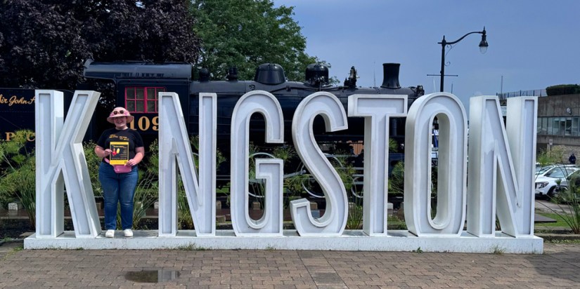 Kingston artist Portia Po Chapman standing as the letter I in the large white Kingston waterfront sign while holding a Being Seen exhibition poster in front of the historic steam engine.