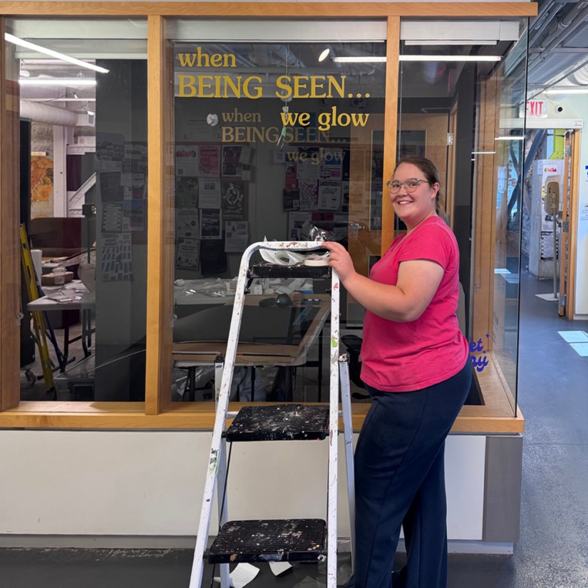 Kingston artist Portia Po Chapman standing beside a stepladder in the Tett Centre hallway, smiling proudly next to the freshly applied gold vinyl title decal "when Being Seen... we glow" on the gallery window.