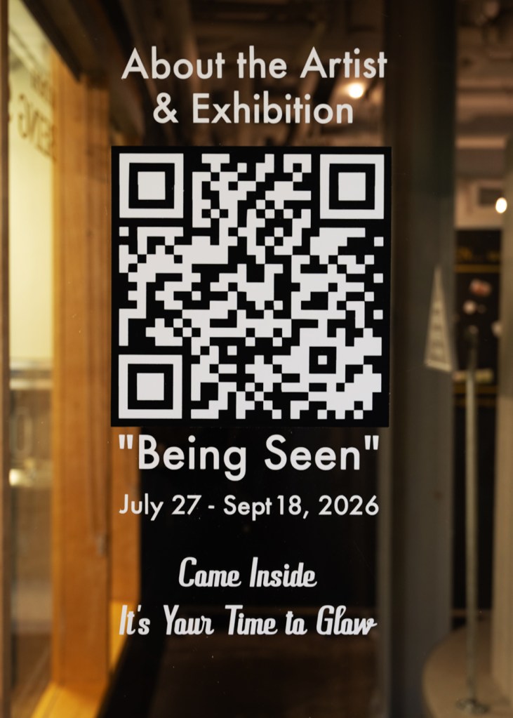 Close-up view of a scannable QR code decal on the Pocket Gallery window at the Tett Centre, titled "About the Artist & Exhibition 'Being Seen'" with dates July 27 โ Sept 18, 2026, and the invitation "Come Inside It's Your Time to Glow" by Kingston artist Portia Po Chapman.