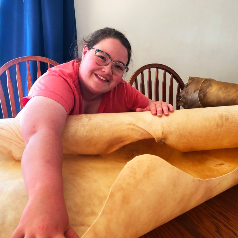 Visual artist Portia Po Chapman leaning over a large, unrolled natural elk rawhide on a flat wooden workspace in her studio, preparing materials for custom frame drum cutting and installation features