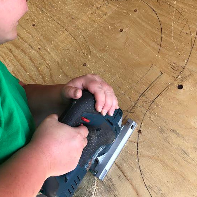 Kingston artist Portia Po Chapman holding a power jigsaw to rough cut along pencil guidelines on a plywood sheet in her studio.