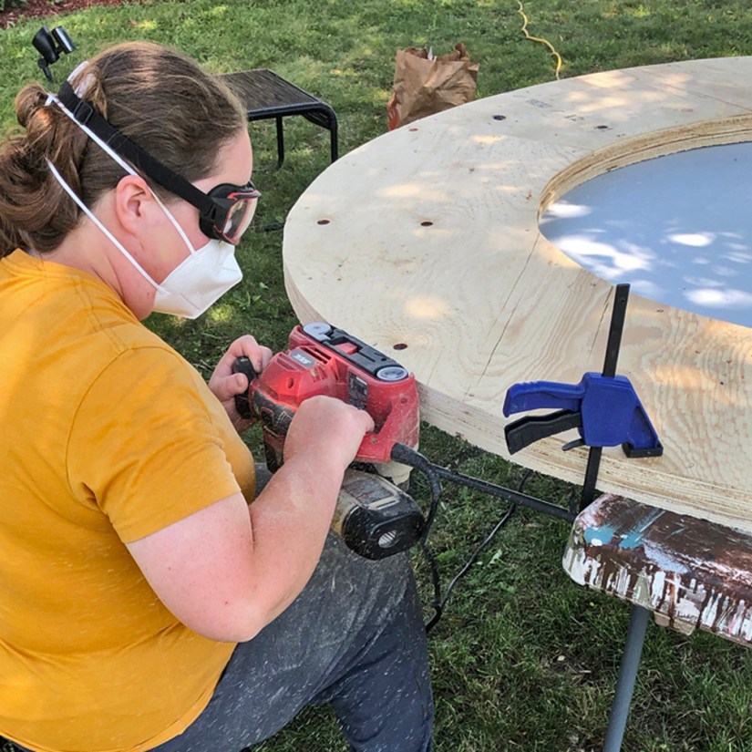 Artist Portia Po Chapman wearing protective mask and goggles while using a power sander on a clamped plywood base under a tree outdoors.
