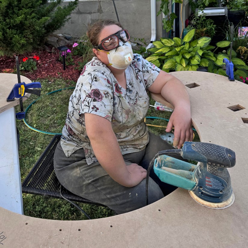 Kingston artist Portia Po Chapman in safety goggles and a dust mask taking a break while seated inside a curved, sanded plinth top section outside.