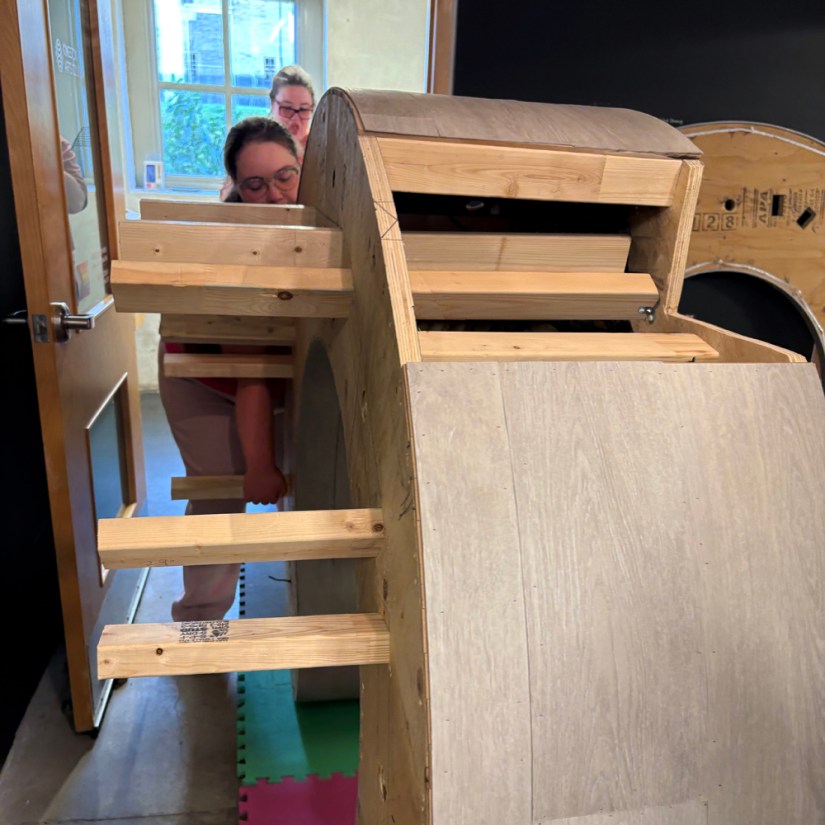 Kingston artist Po Chapman and her assistant maneuvering the upright Glow Chamber wooden plinth base inside the KAC Pocket Gallery doorway, with the top panel leaning against the gallery wall in the background.