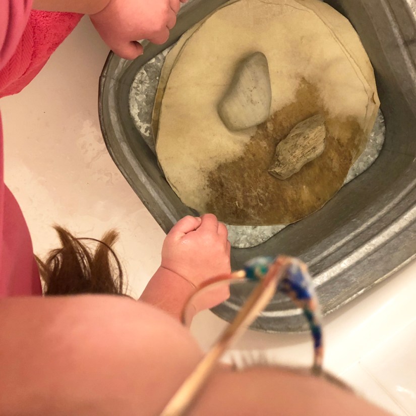Top-down view of rawhide circles submerged in water inside a vintage galvanized washtub, held down by river rocks during a studio soaking process by Portia Po Chapman