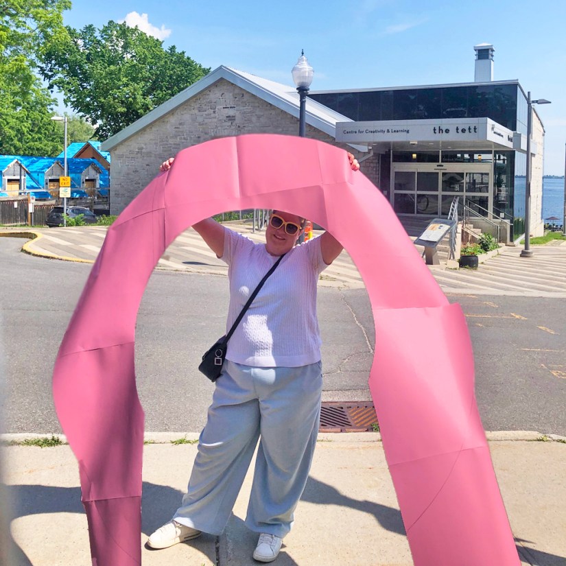 Kingston artist Portia Po Chapman holding a large pink paper arch template in front of the Tett Centre for Creativity and Learning.