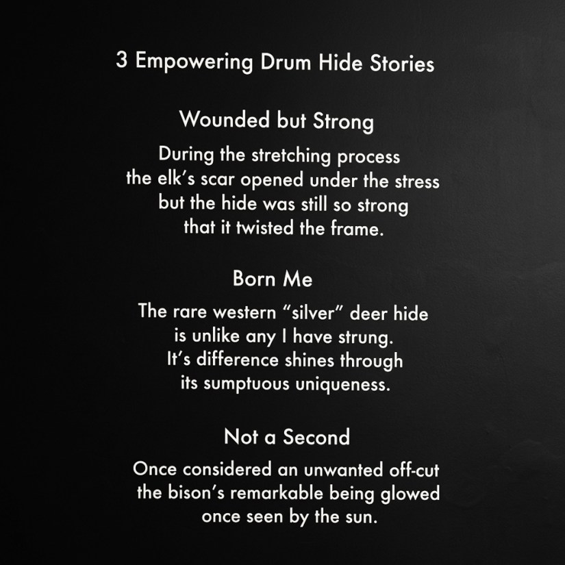 Close-up view of the black didactic wall graphic panel titled "3 Empowering Drum Hide Stories" by Kingston artist Portia Po Chapman, describing the origin stories for her featured glowing rawhide drums: Wounded but Strong, Born Me, and Not a Second.