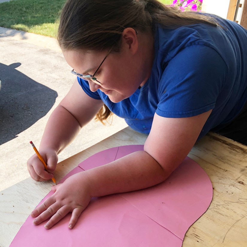 Kingston artist Portia Po Chapman leaning over a wooden workbench in her studio tracing a pink paper template onto plywood with a pencil.