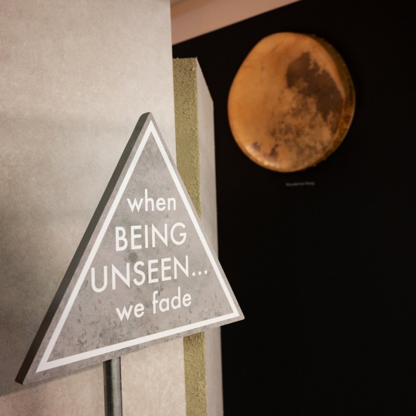 A grey triangular caution sign reading "when Being Unseen... we fade" positioned outside the Glow Chamber installation by Kingston artist Portia Po Chapman, with a glowing golden elk rawhide drum mounted on the dark wall in the background.