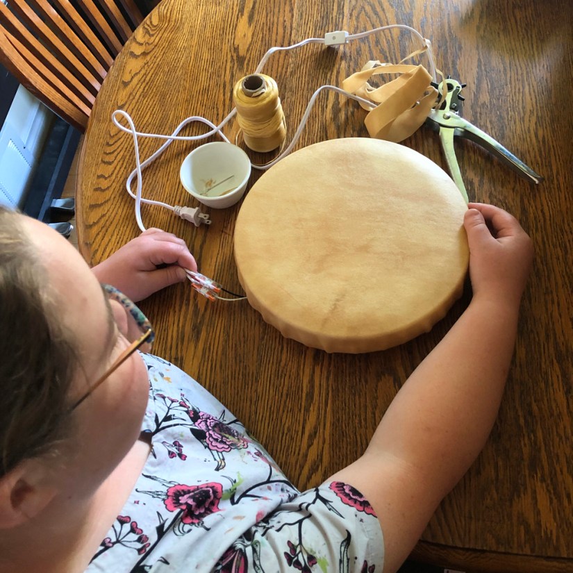 Top-down view of Kingston artist Portia Po Chapman holding orange Wago Lever-Nut connectors in her left hand as she connects a lamp cord to a smooth rawhide-wrapped LED vessel sitting on a wooden studio table surrounded by tools.